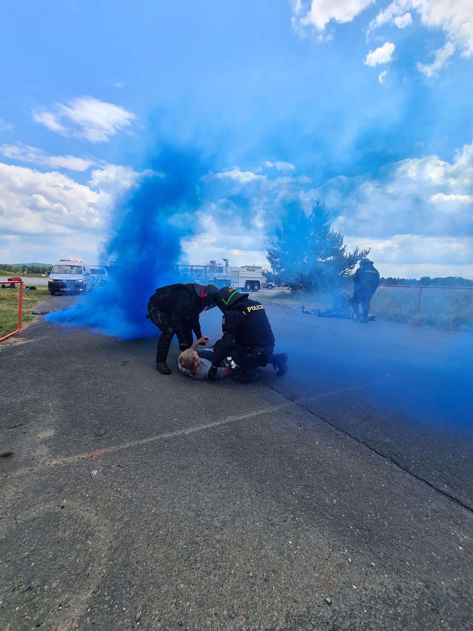 Policisté v zásahové výstroji drží osobu ležící na zemi uprostřed modrého dýmu při výcvikovém cvičení.